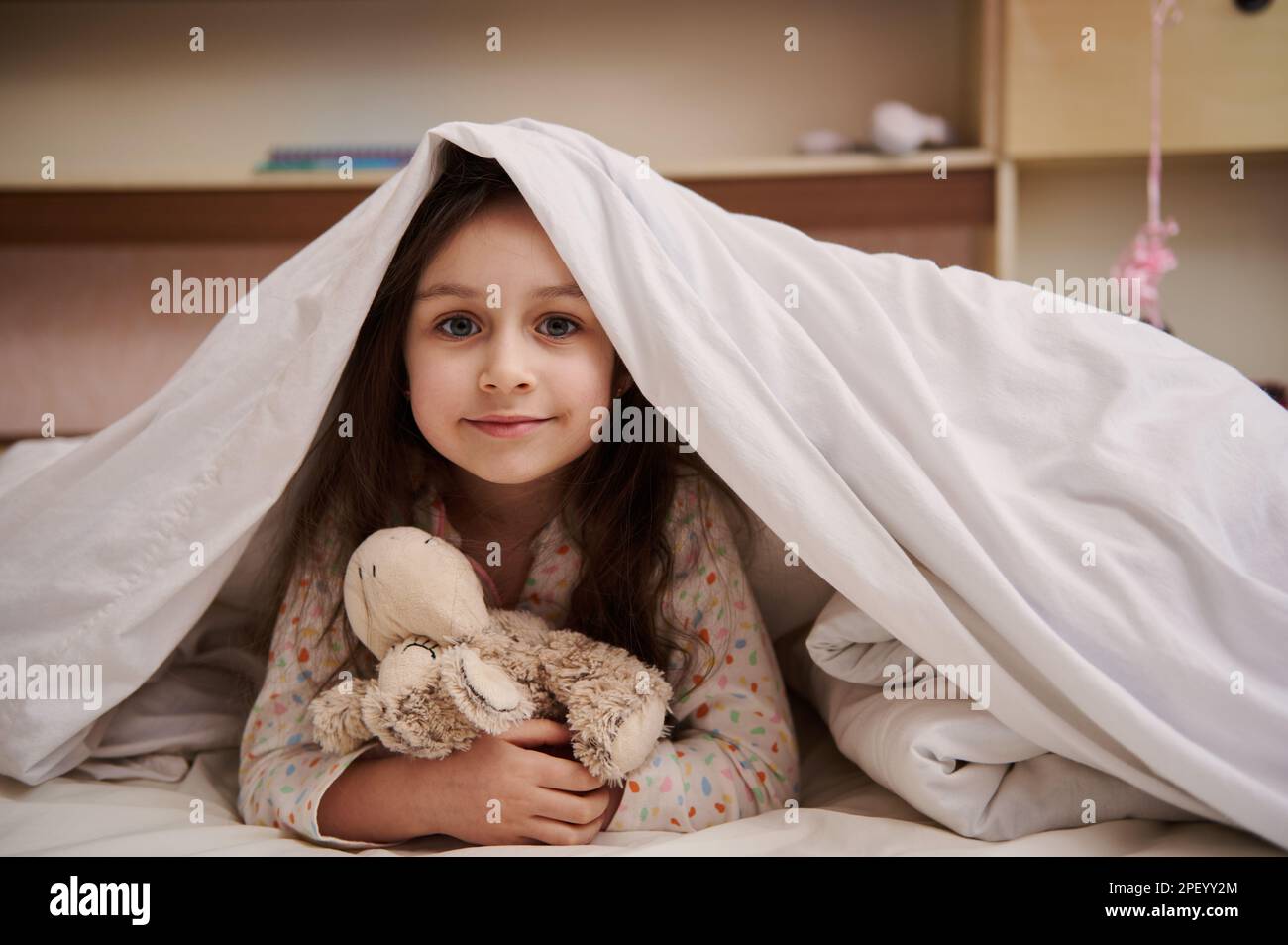 Bella bambina in pigiama, abbracciando la sua pecora peluche giocattolo, sdraiata sul letto sotto il bianco soffice comfodtrabile Foto Stock