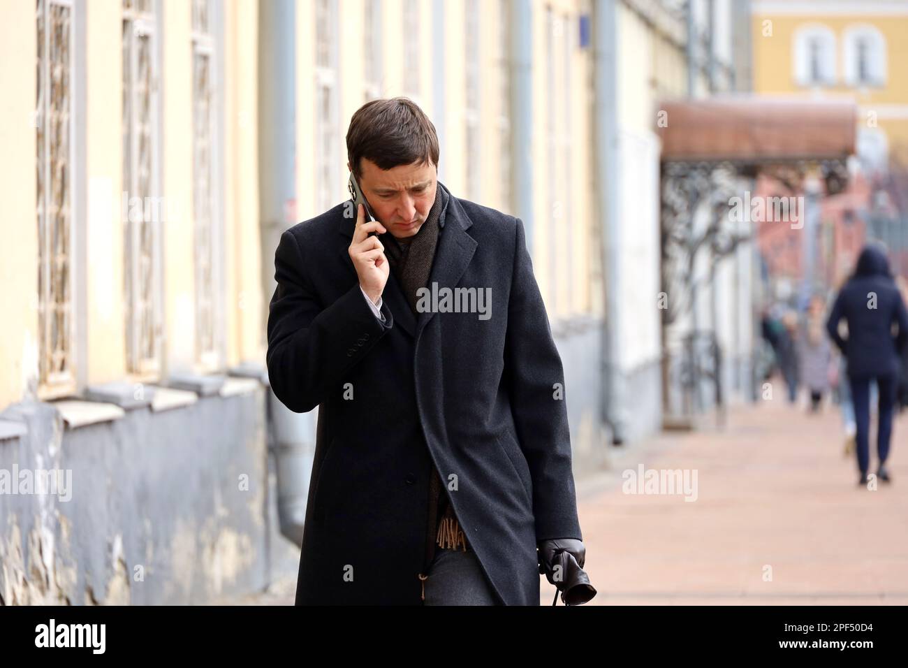 Un uomo sconvolto in un cappotto che parla sullo smartphone mentre cammina per strada. Uso del telefono cellulare nella città primaverile Foto Stock