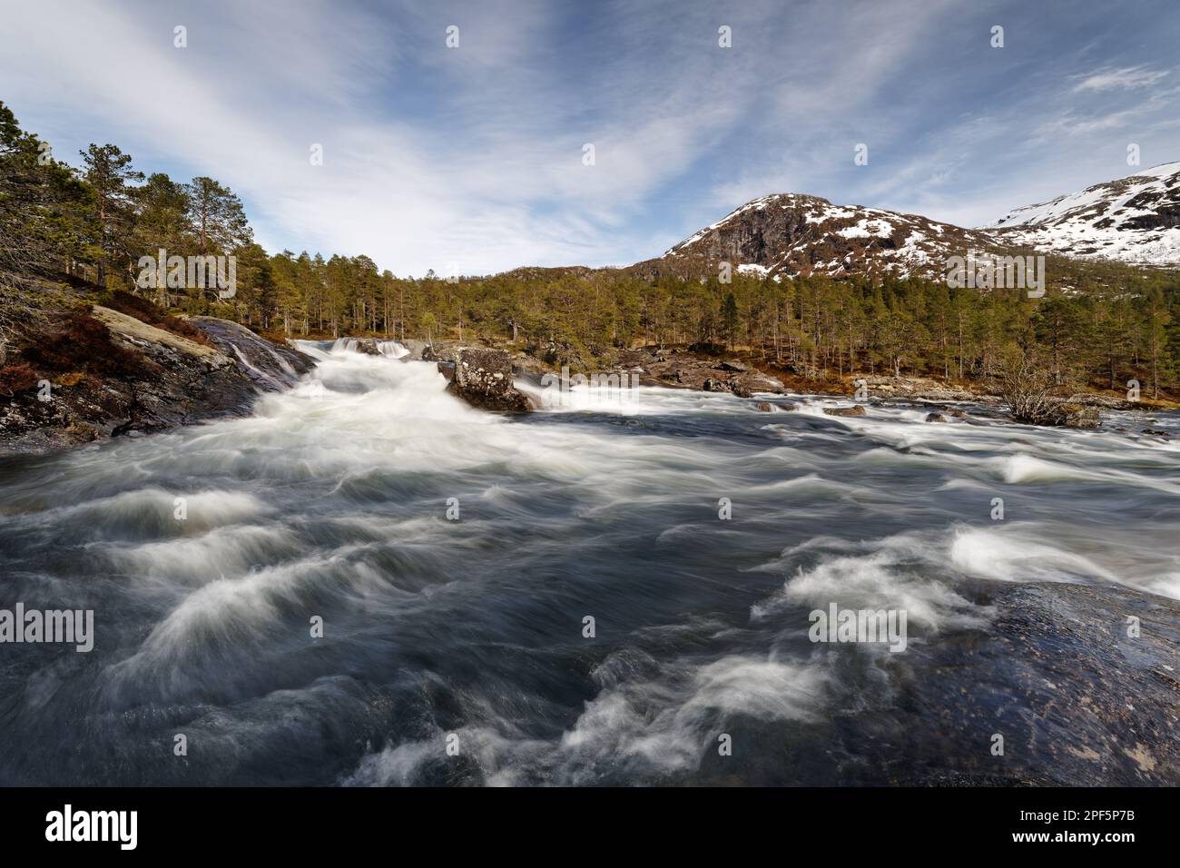 Vista su un fiume con grandi e piccole rapide, il sole splana in acqua, foresta e montagne parzialmente innevate sullo sfondo, girato con lungo Foto Stock