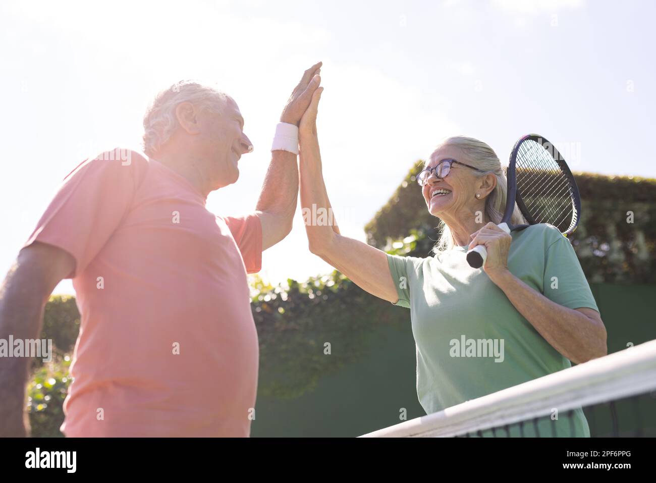Amici anziani caucasici felici con racchette da tennis alta fiving al campo da tennis nella giornata di sole Foto Stock