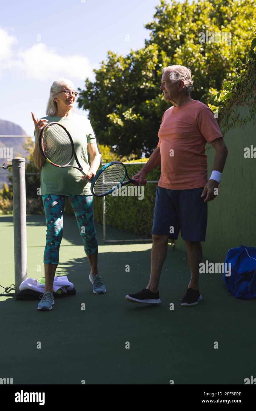 Amici anziani caucasici felici con racchette da tennis che parlano al campo da tennis nella giornata di sole Foto Stock