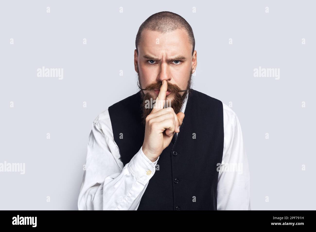 Mantieni il silenzio. Ritratto di un uomo serio e rigoroso con barba e baffi in piedi e con un gesto shh, guardando la fotocamera con un'espressione bossy. Studio in interni isolato su sfondo grigio. Foto Stock