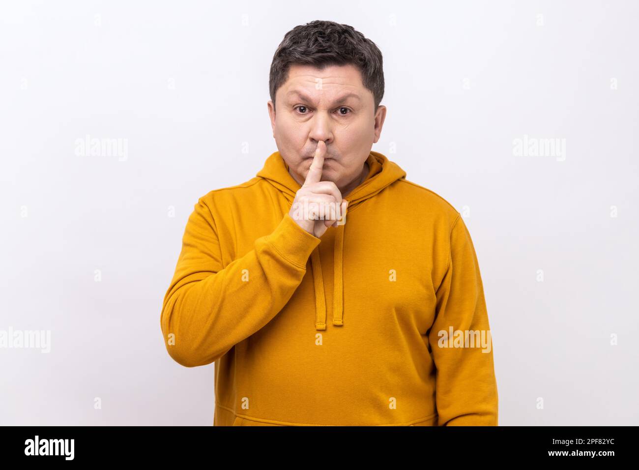 Ritratto di un uomo di mezza età che tiene il dito sulle labbra facendo un gesto di silenzio, chiedendo di tenere segreto, non parlare, indossando felpa con cappuccio in stile urbano. Studio in interni isolato su sfondo bianco. Foto Stock