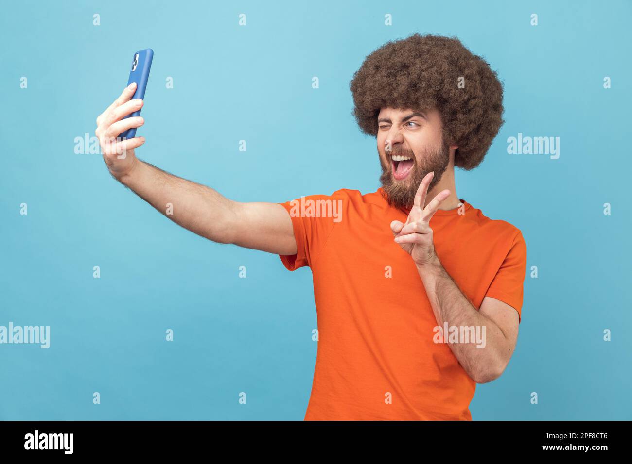 Ritratto dell'uomo con acconciatura afro che indossa una T-shirt arancione con un gesto v, che si posa alla fotocamera del telefono, fa selfie o fa videochiamata. Studio in interni isolato su sfondo blu. Foto Stock