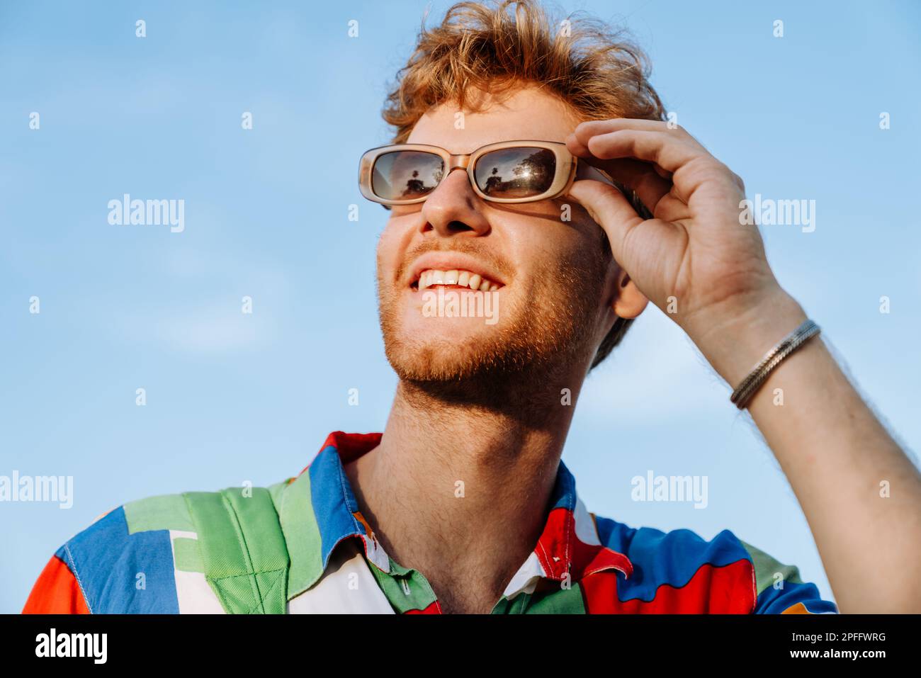 Ritratto di un giovane bel giovane uomo sorridente alla moda in camicia colorata che regola i suoi occhiali da sole e guarda da parte Foto Stock