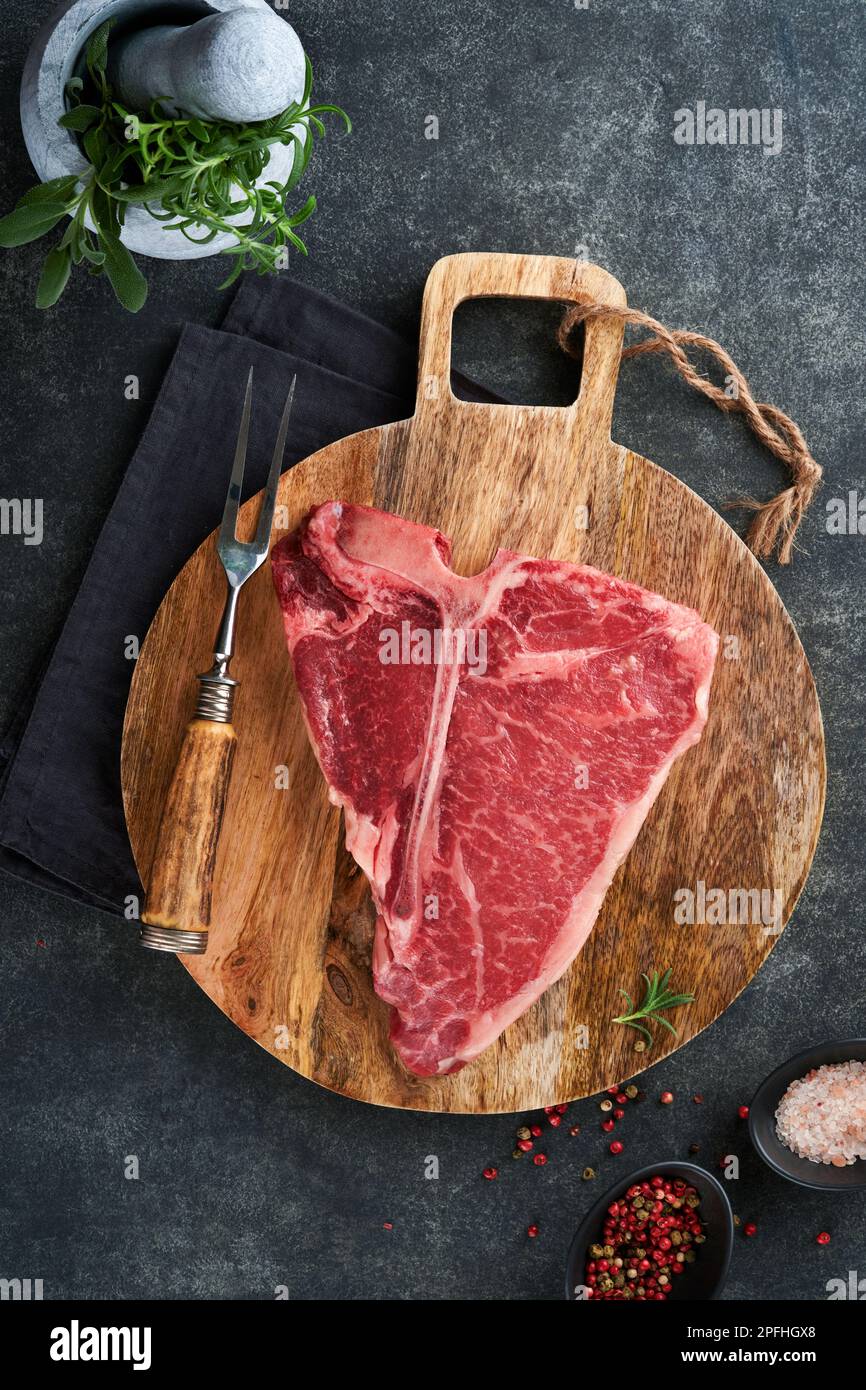 Bistecca con osso a T crudo spesso. Carne di manzo cruda e stagionata bistecca di manzo su cinghiale da taglio con erbe e sale su fondo scuro. Vista dall'alto e copia Foto Stock