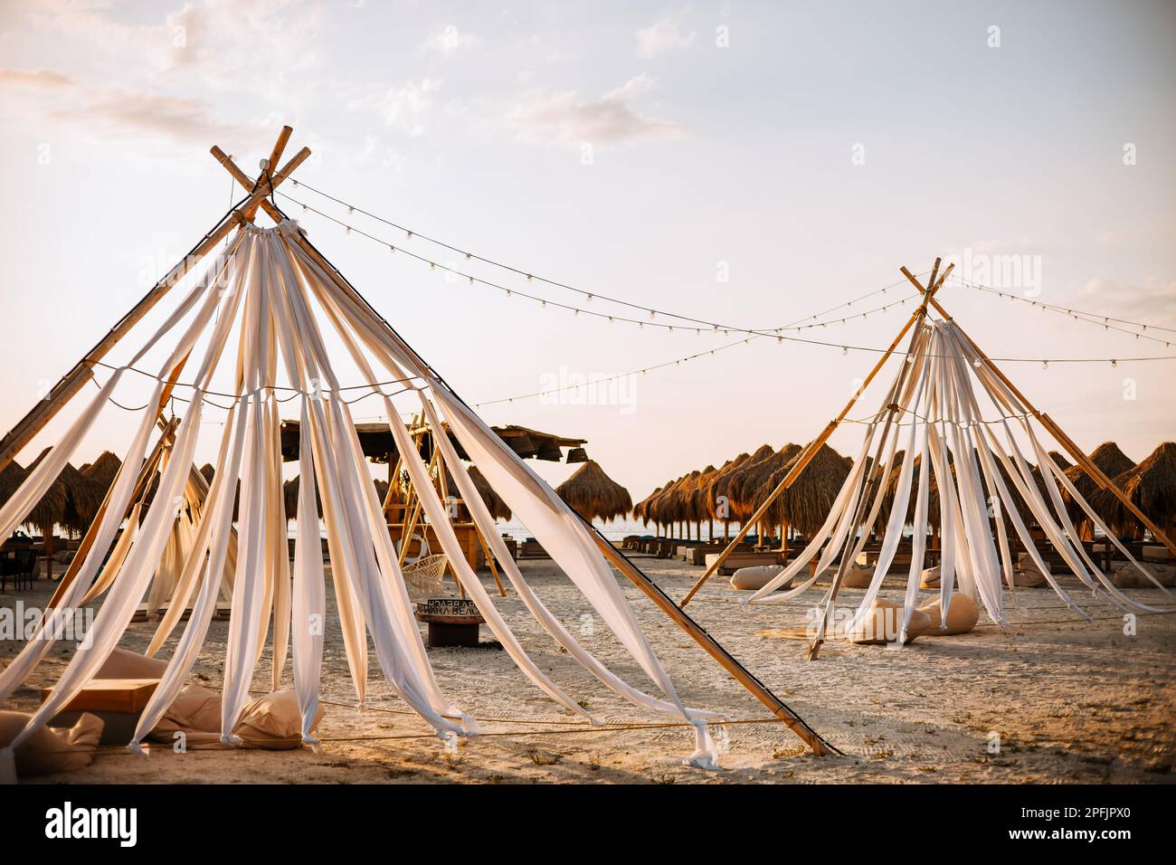 Tende boho in barrette di legno e materiale bianco sulla spiaggia. Sullo sfondo, vicino al mare, si possono vedere ombrelloni di paglia e lettini Foto Stock