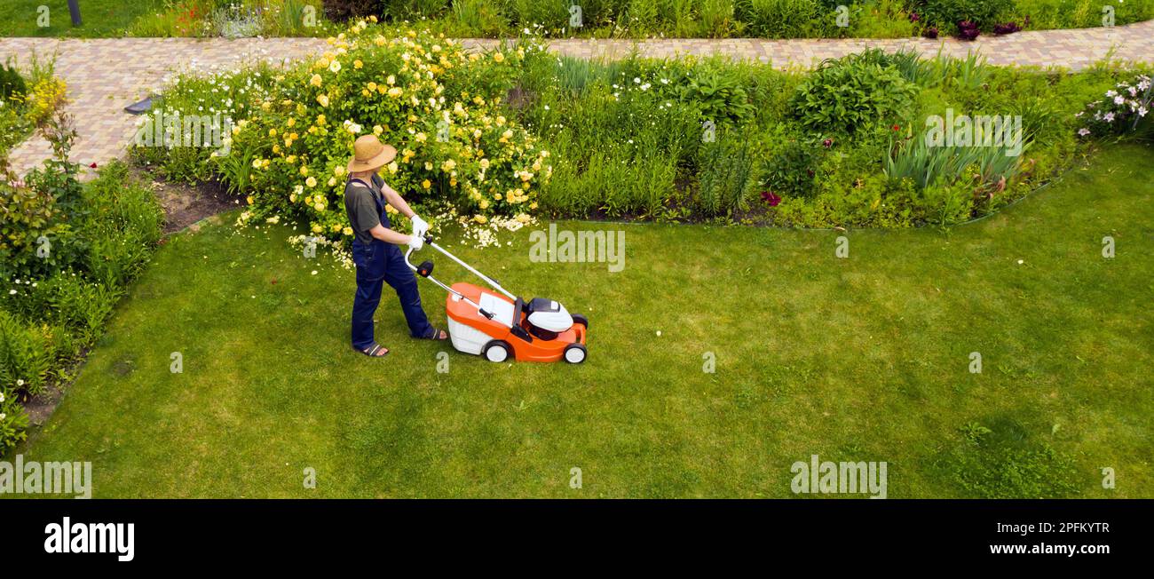 il giovane uomo sta falciando un prato con un rasaerba nel suo verde giardino estivo floreale. Foto Stock