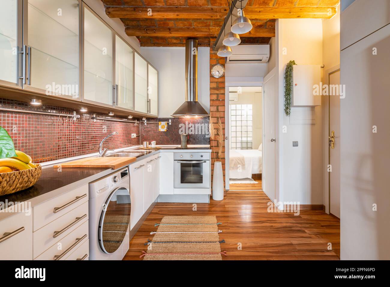 Cucina elegante e accogliente con elettrodomestici moderni e soffitto in legno con soppalco che si affaccia sulla camera da letto aperta con parete in mattoni di vetro. Concetto di design laconico Foto Stock