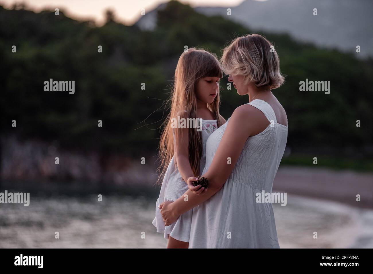 Primo piano ritratto di madre incinta con bambina che cammina mano in mano in abiti bianchi al tramonto sul mare. Assistenza ai bambini. Maternità sana. Foto Stock