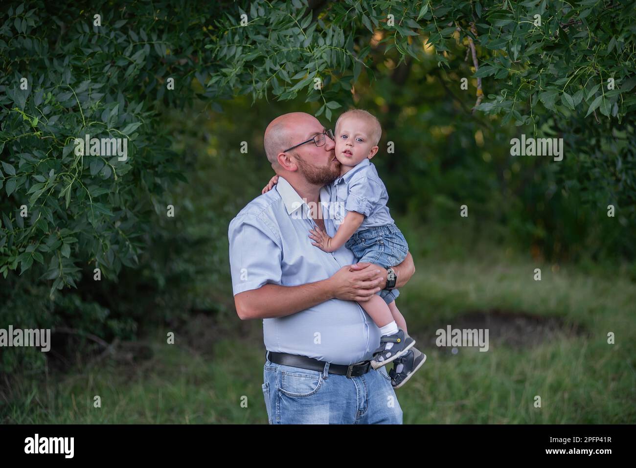 L'uomo calvo in occhiali tiene il bambino in braccio nel parco. Padre in jeans abbraccia il figlio nella natura al di fuori della città. Il ragazzino ride, divertendosi con il papà su un Foto Stock