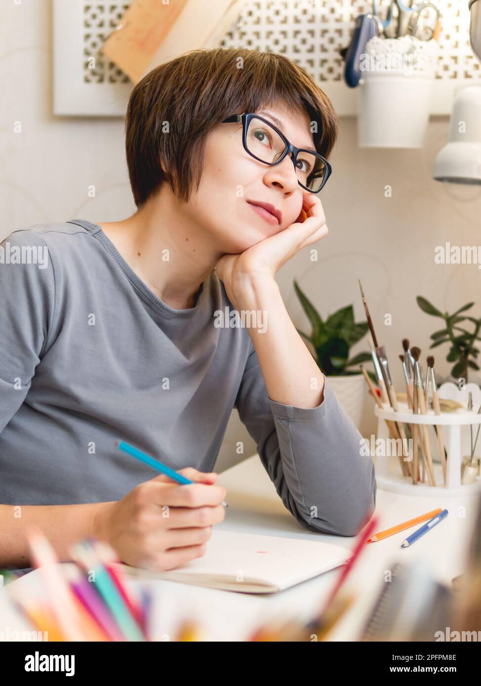 Donna con taglio corto dei capelli sta disegnando nel taccuino. Hobby calmante, tempo libero antistress. Artista al lavoro. Ambiente di lavoro accogliente. Foto Stock