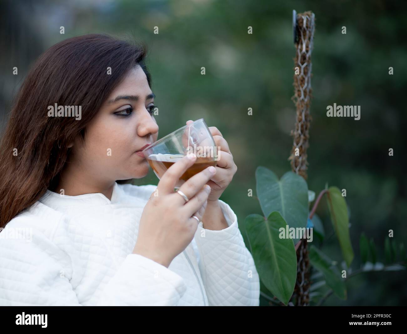 Le giovani donne che tengono una tazza da tè trasparente con entrambe le mani bevono tè nero in un giardino indossando una giacca di lana bianca Foto Stock