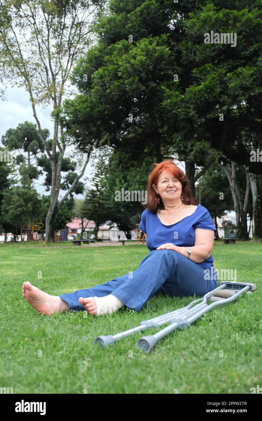 Donna matura che riposa sull'erba, sorridente, a piedi nudi, con una fasciatura sul piede sinistro e stampelle al suo fianco. Concetti: Atteggiamento positivo verso un Foto Stock