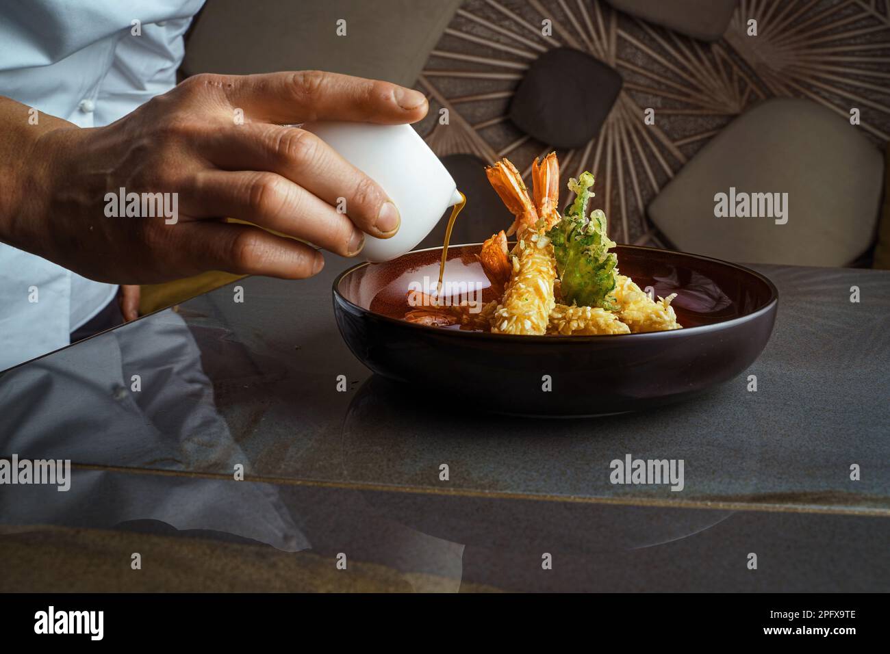 Vista ravvicinata di un'elegante ciotola nera piena di gamberi tempura e verdure, sormontata da una salsa agrodolce e dolce versata dalla mano dello chef Foto Stock