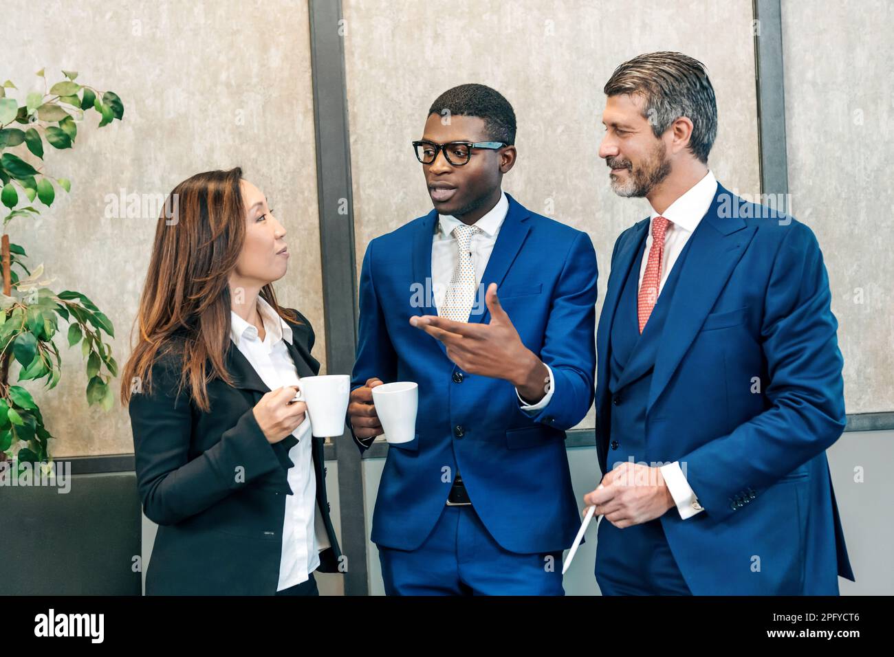 Colleghi multietnici in abiti di classe in piedi con tazze di bevande calde e parlare mentre si è in pausa dal lavoro Foto Stock