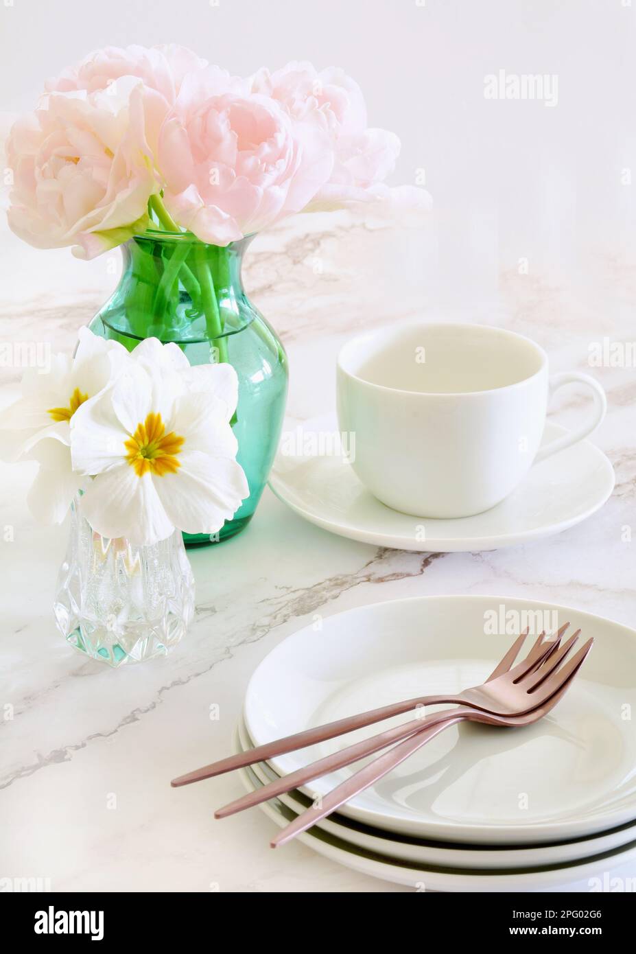 Delicati tulipani rosa con fiori di primula bianchi e piatti da dessert con forchette in oro rosa su fondo in marmo. Formato verticale con focalizzazione sui tulipani. Foto Stock