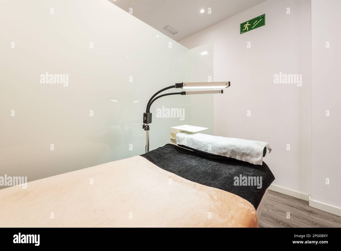 Cabina per trattamenti estetici con pareti in vetro temperato con tavolo massaggiante, asciugamani neri e pulizia con doppie lampade pieghevoli e pavimento in laminato Foto Stock