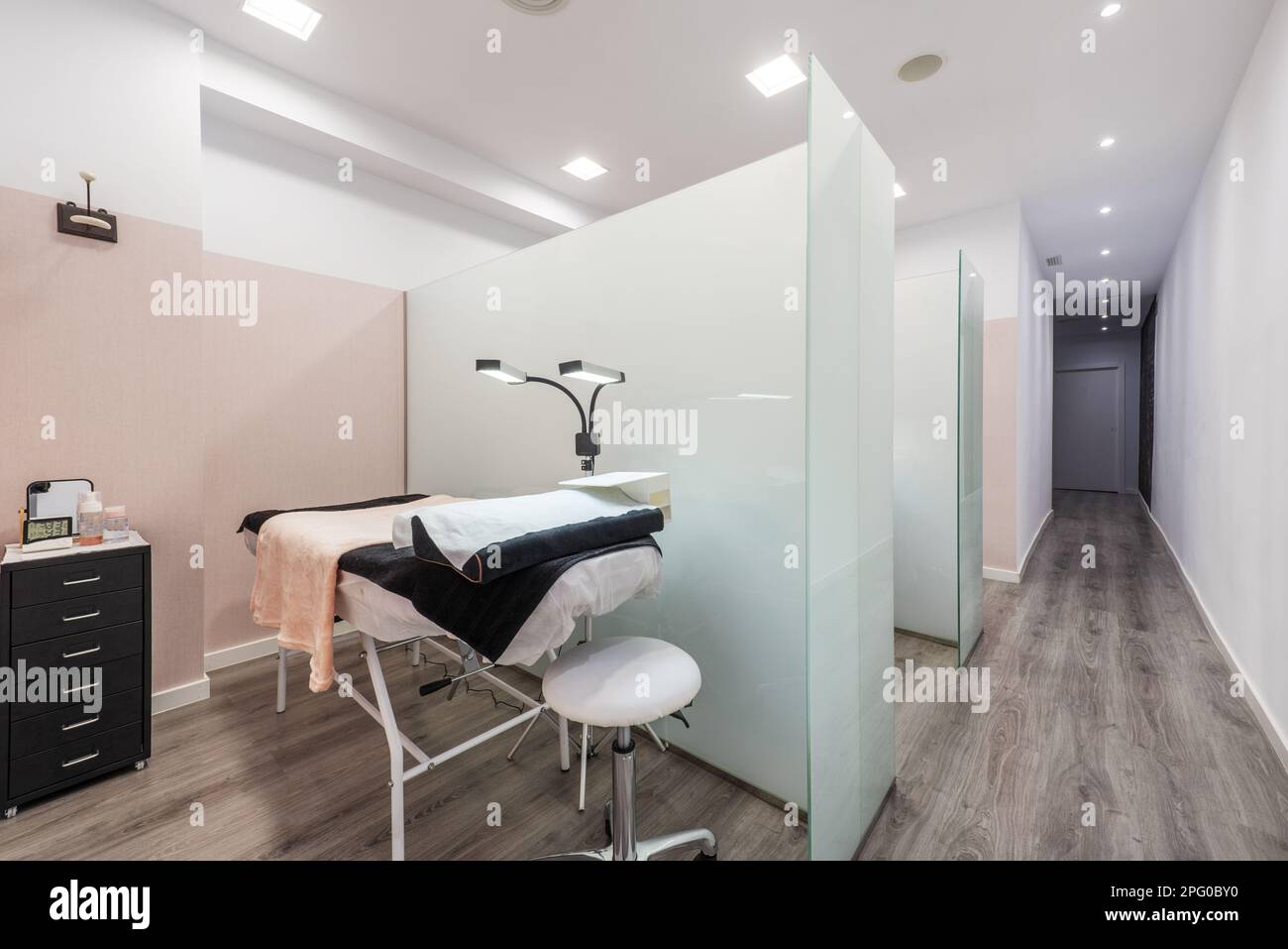 Un corridoio con diverse cabine di trattamento estetico con pareti in vetro temperato con tavoli da massaggio, asciugamani neri e pulizia con doppie lampade pieghevoli, Foto Stock