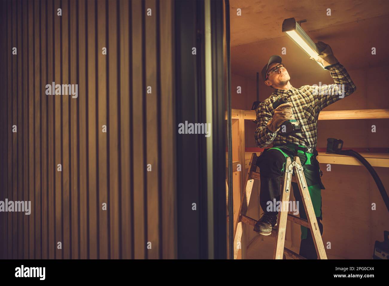 Caucasian Electrician in His 40s Installazione del dispositivo di luce a LED nel soffitto di Shed. Piccola illuminazione di edifici in legno Foto Stock