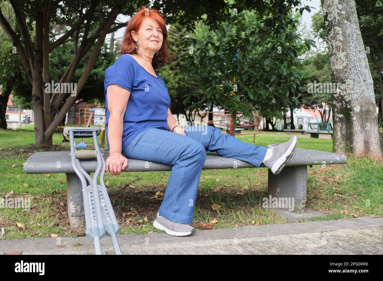 Donna matura ispanica con una ferita del piede si siede su una panca del parco con un paio di stampelle propped in su vicino a lei. Concetti: Atteggiamento positivo verso l'adve Foto Stock