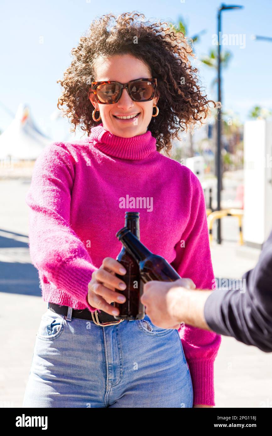 Capelli ricci giovane donna caucasica fare un brindisi con un coltivatore in strada tenendo due bottiglie di birra in una giornata di sole. Foto Stock