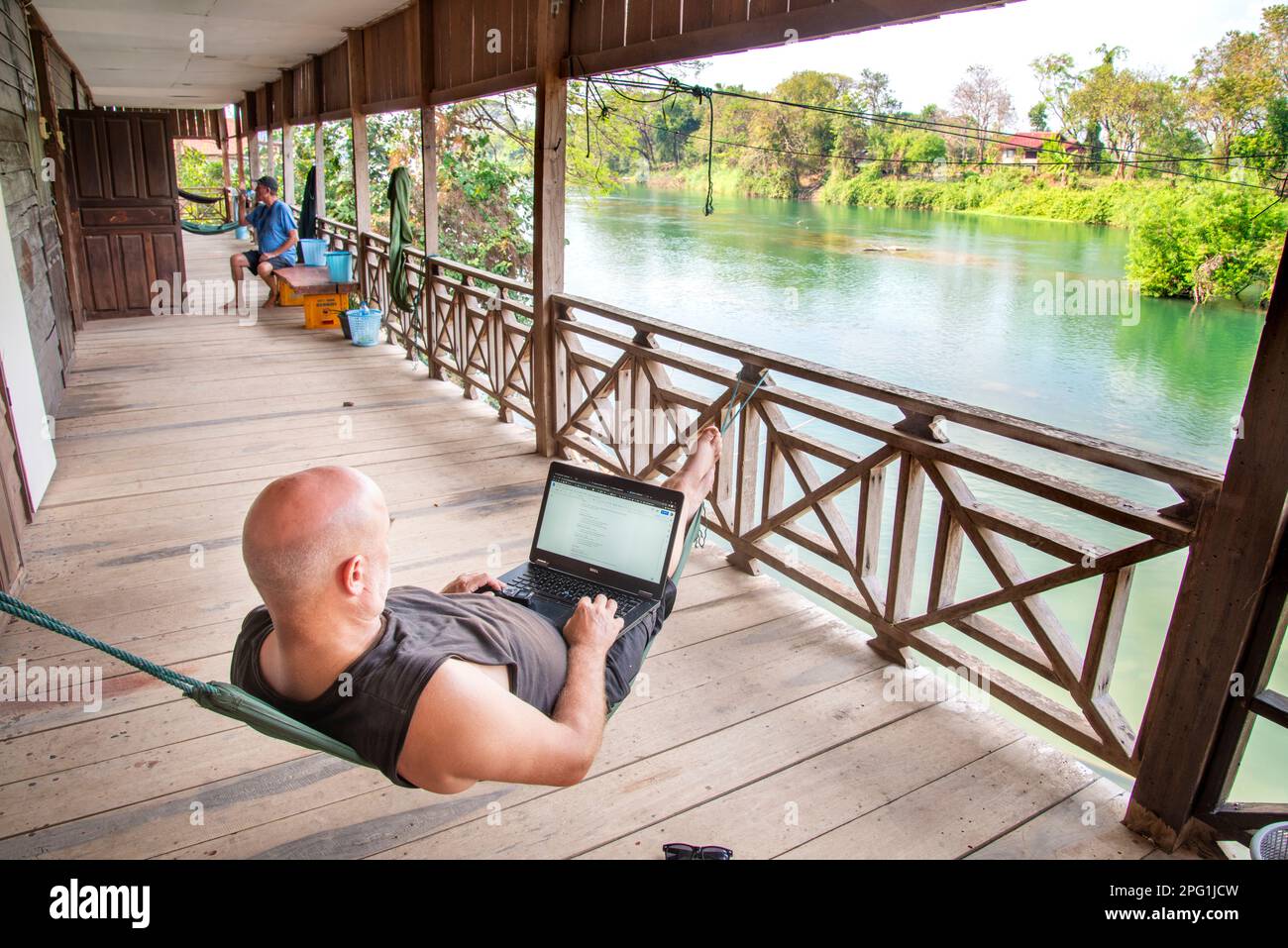 Lavorando a distanza sul suo computer in si Phan Don, Laos meridionale, mentre si viaggia per il mondo, rilassarsi su un balcone pensione in legno che si affaccia sul Mekon Foto Stock