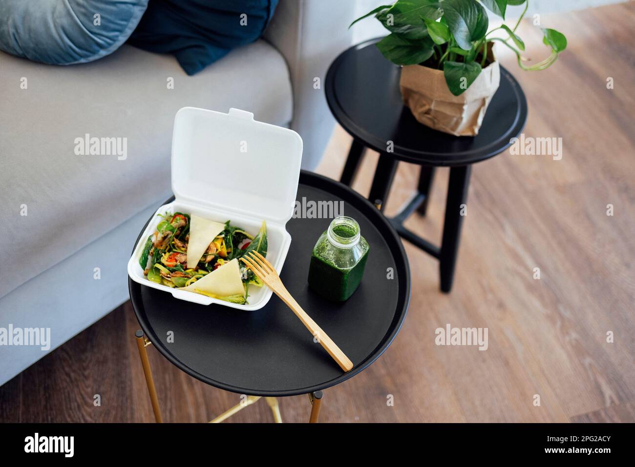 Primo piano del cibo da portare via in contenitore bianco con forchetta di legno e bottiglia plactica di frullato verde. Il pasto si trova su un tavolo da caffè nero rotondo. Legno b Foto Stock