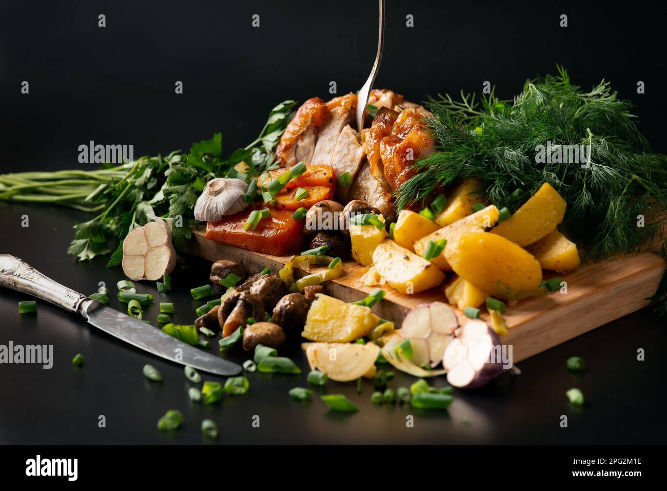 Pollo fritto con patate e funghi su fondo scuro. Pollame affumicato con verdure e forchetta d'annata. Pollo arrosto tagliato a pezzi. Foto Stock