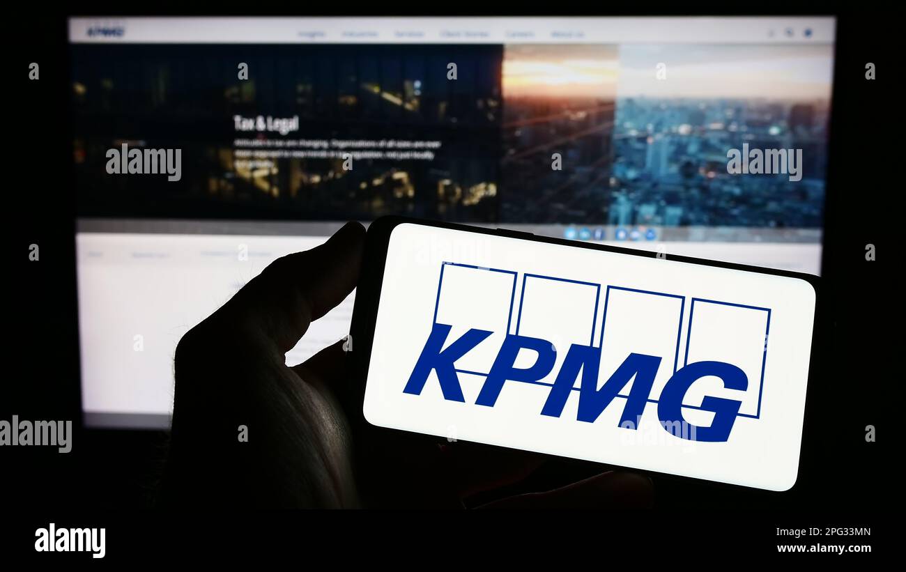 Persona che tiene il telefono cellulare con il logo della società di servizi professionali KPMG sullo schermo di fronte alla pagina web aziendale. Messa a fuoco sul display del telefono. Foto Stock