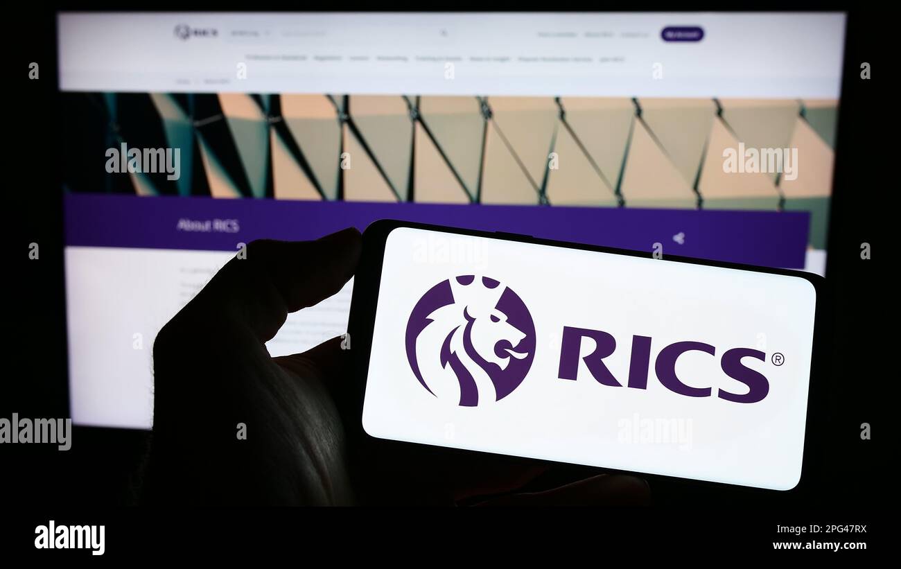 Persona che tiene un telefono cellulare con il logo della Royal Institution of Chartered Surveyors (RICS) sullo schermo davanti alla pagina web. Messa a fuoco sul display del telefono. Foto Stock
