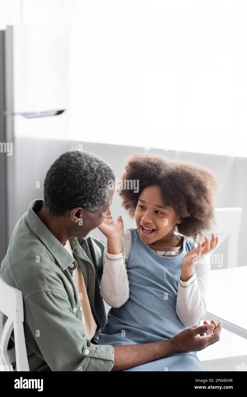 ragazza afro-americana eccitata con capelli ricci gesturing mentre parlando con nonno allegro a casa, immagine di scorta Foto Stock