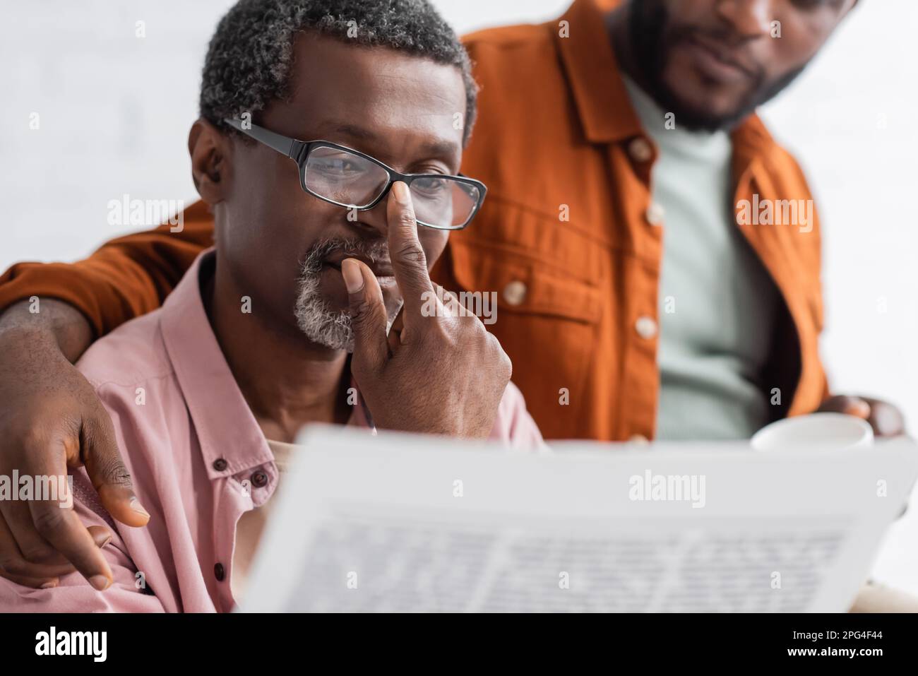 Uomo africano-americano maturo in eyeglasses che legge il giornale vicino al figlio sfocato nel paese, immagine di scorta Foto Stock