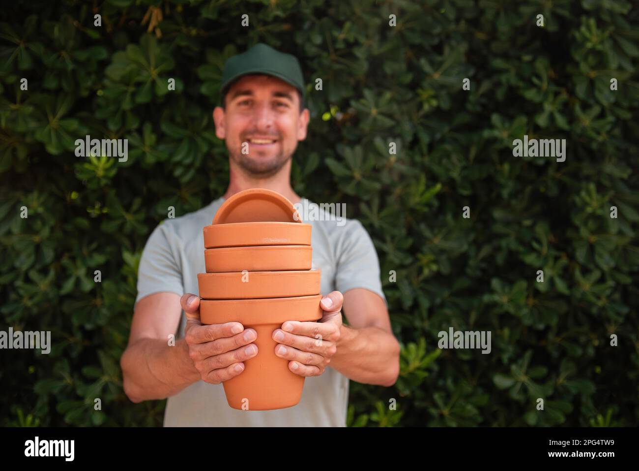 Primo piano ritratto uomo giardiniere in berretto verde tiene in mano pentole di terracotta terracotta contro lo sfondo di un recinto sempreverde. Giardinaggio, piccola impresa, Foto Stock