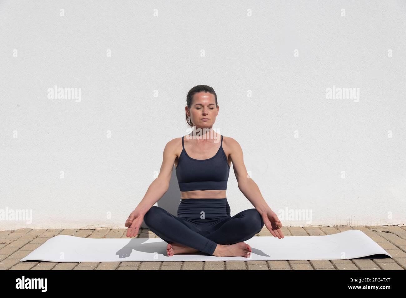 Donna seduta su un tappetino yoga meditando fuori contro una parete bianca al sole Foto Stock