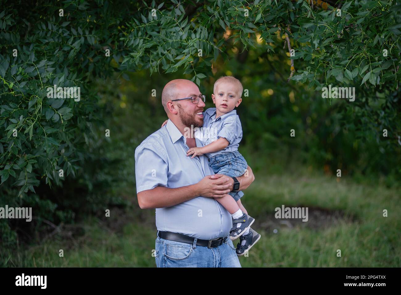 L'uomo calvo in occhiali tiene il bambino in braccio nel parco. Padre in jeans abbraccia il figlio nella natura al di fuori della città. Il ragazzino ride, divertendosi con il papà su un Foto Stock