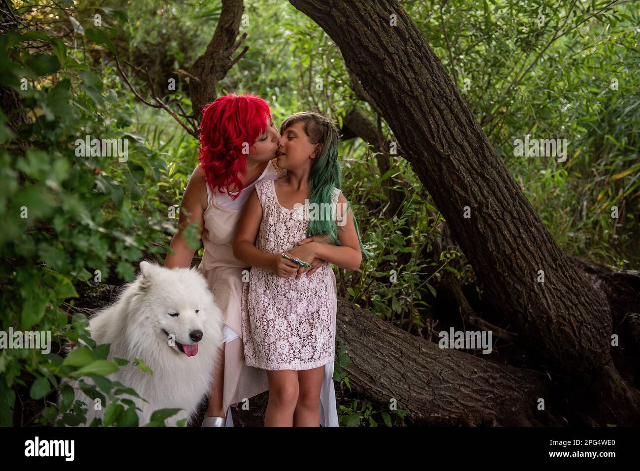 Nella Foresta Verde, da piccolo ruscello diversità, madre con capelli rosa abbraccia figlia. Il bianco soffice samoyed custodisce la famiglia. In viaggio con animali domestici Foto Stock