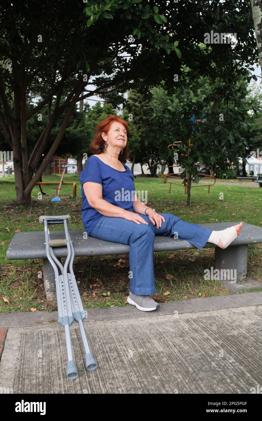 Donna matura latina con una ferita seduta su una panca del parco con una bendatura sul suo piede sinistro e stampelle propped in su vicino a lei. Concetti: Atti positivi Foto Stock