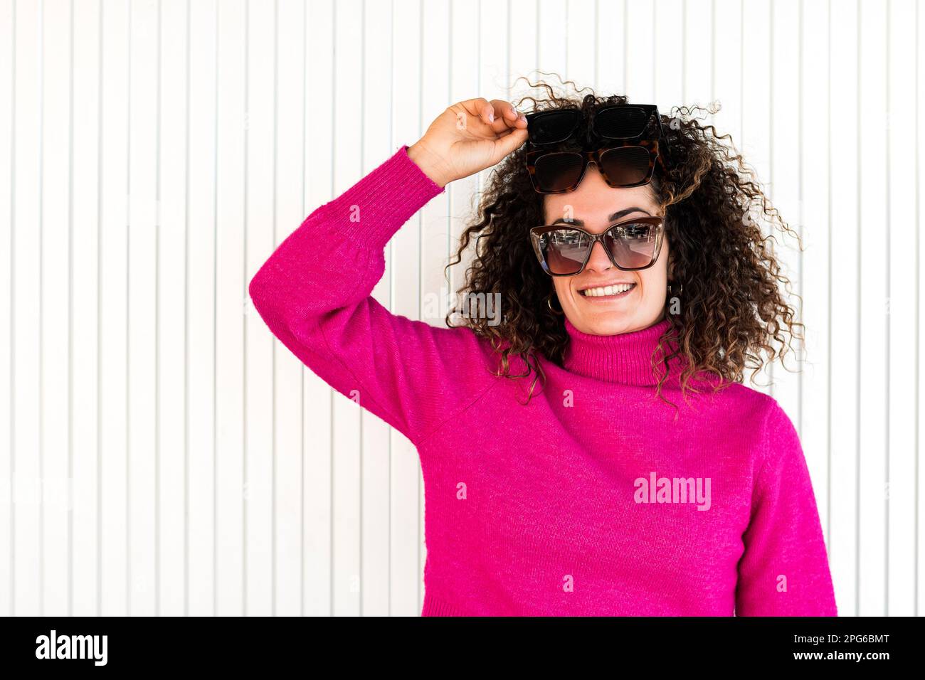 Donna adulta sorridente con capelli castani ricci in occhiali da sole alla moda e maglione rosa in piedi vicino alla parete a strisce e alla testa a contatto Foto Stock