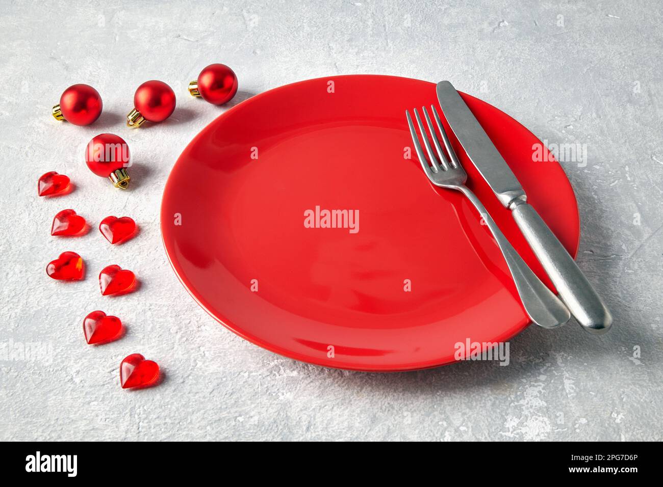 Piatto di ceramica rosso vuoto con posate circondate da palline rosse di Natale e cuori su un tavolo di cemento chiaro. Mockup di esposizione del cibo Foto Stock