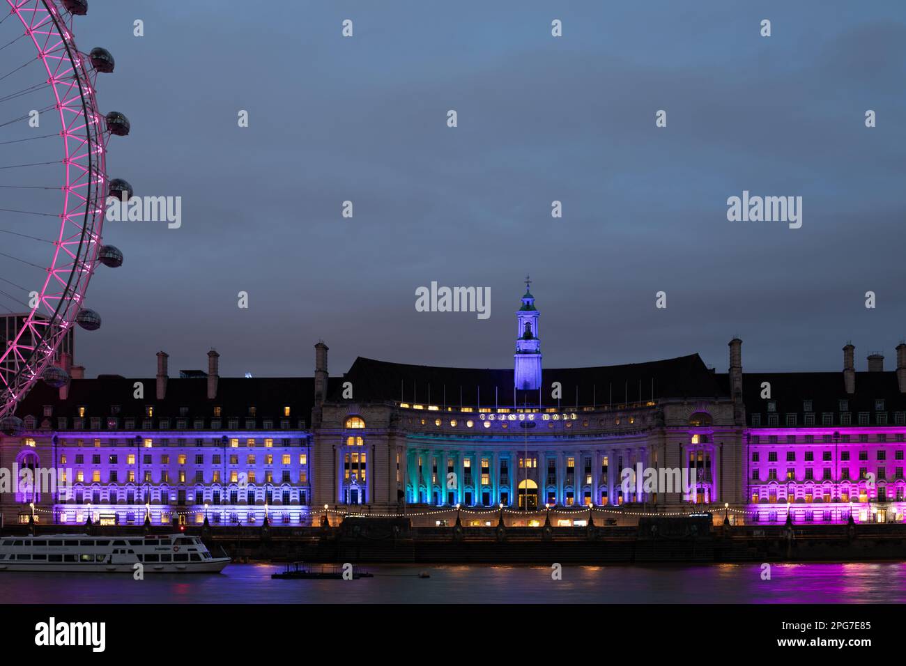 La County Hall, illuminata dai colori viola, blu e rosa, sorge di notte sulla riva sud del Tamigi, accanto al London Eye. Foto Stock