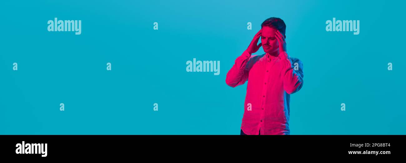 Giovane uomo che soffre di stress e mal di testa, tenendo la testa con le mani, in posa su sfondo blu studio in luce rosa al neon. Foto Stock