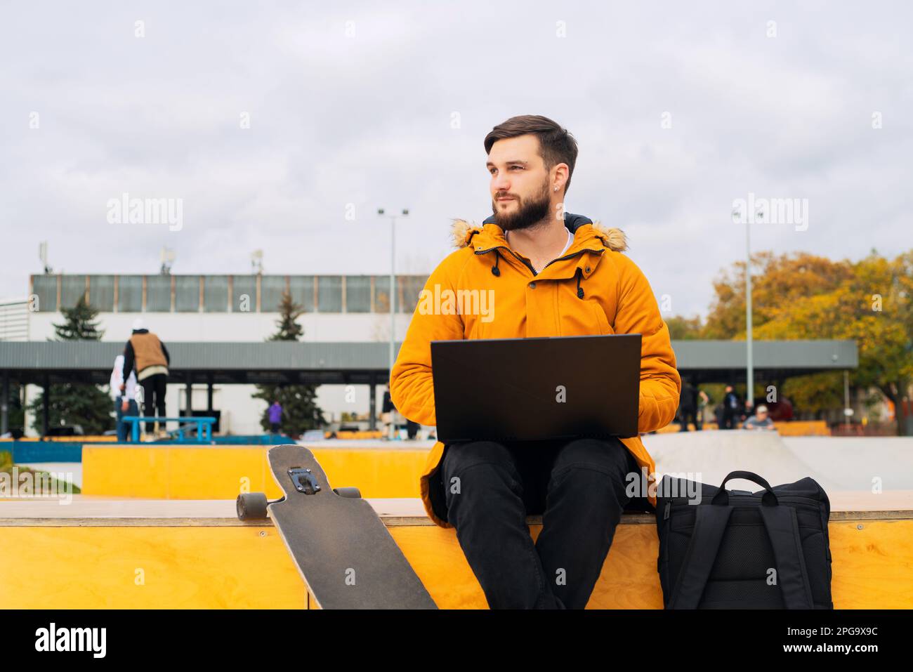 Giovane freelancer casual elegante e alla moda che lavora con un notebook moderno e che usa in uno smartphone mentre si siede su una panca con un longboard all'aperto Foto Stock