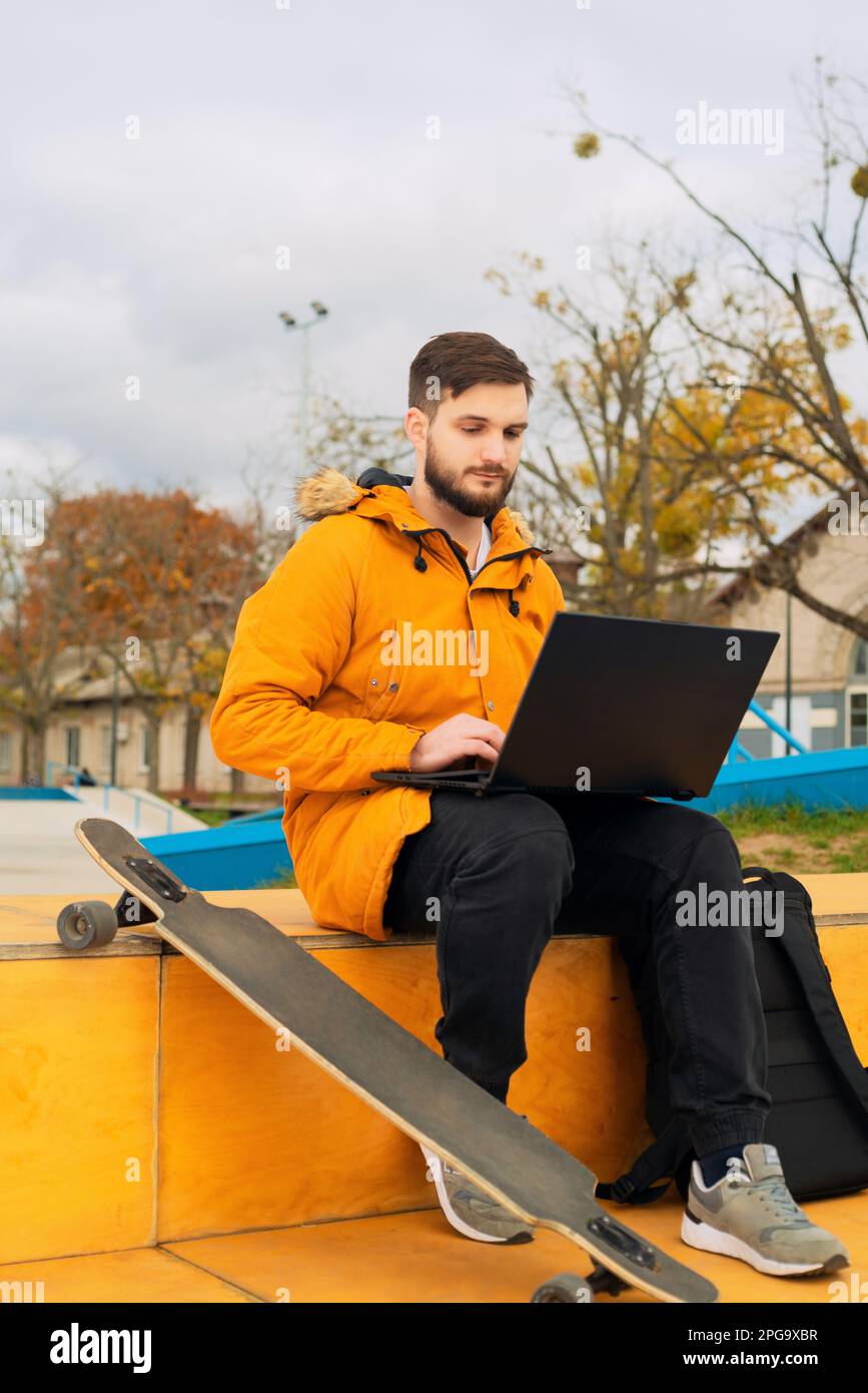 Giovane freelancer casual elegante e alla moda che lavora con un notebook moderno e che usa in uno smartphone mentre si siede su una panca con un longboard all'aperto Foto Stock