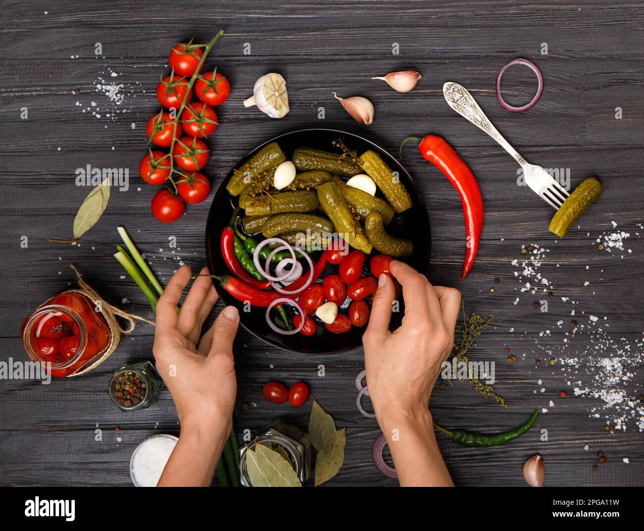 Pomodori ciliegini sottaceto, cetrioli di cetrioli in un piatto nero e le mani della ragazza stendono le verdure su uno sfondo di legno scuro vista dall'alto. Foto Stock