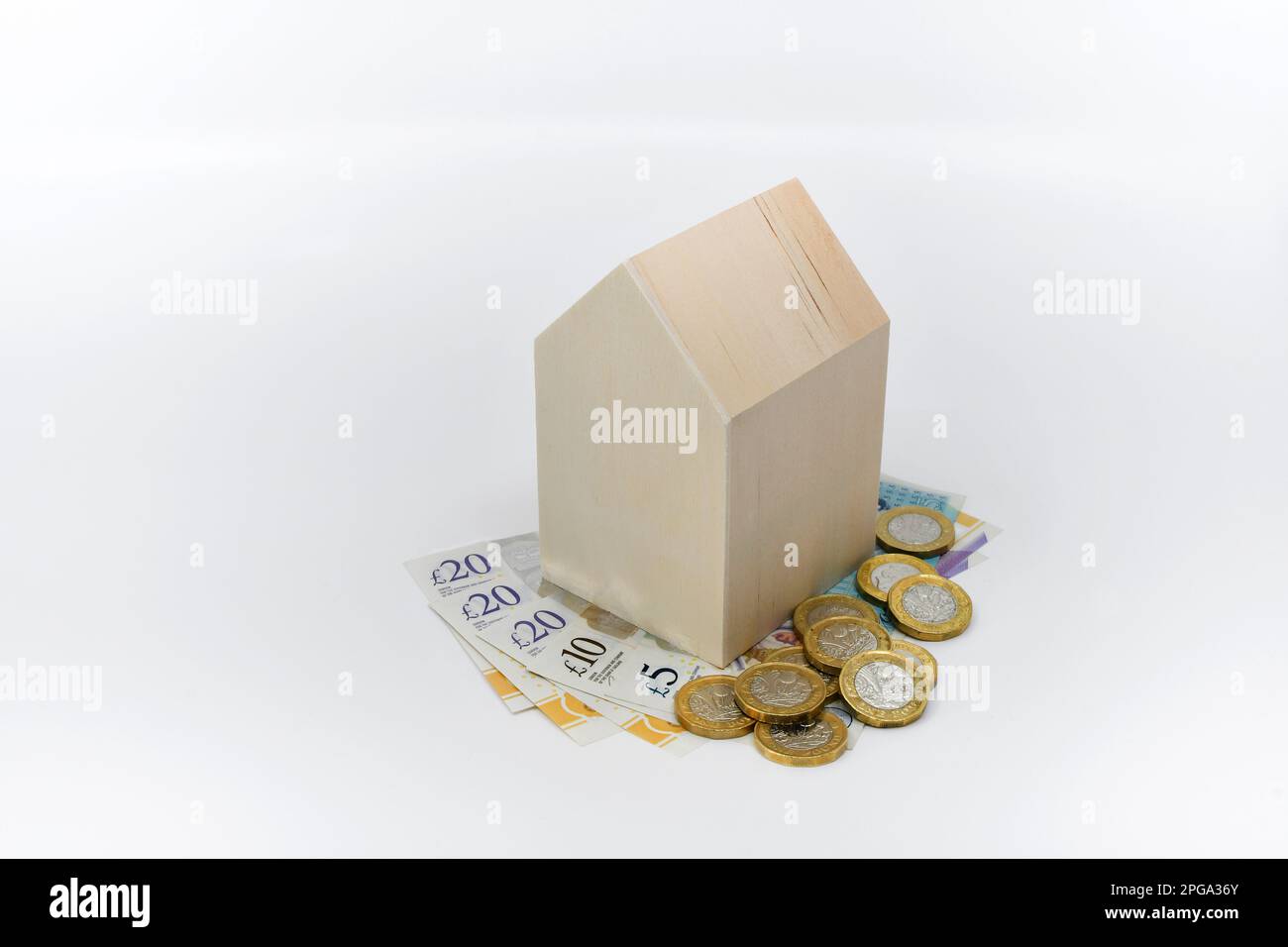 Piccolo modello di casa in legno con banconote e monete britanniche da una libbra su un fondo di wahite semplice. Nessuna gente. Spazio di copia. Costo del concetto di vita. Foto Stock