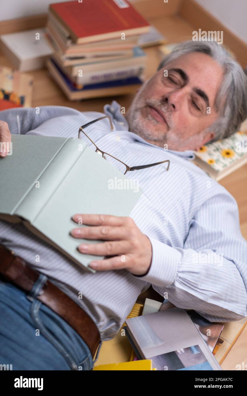 Uomo maturo con barba grizzled e capelli che dormono tra molti libri distribuiti sul pavimento. Addormentarsi mentre si legge con un libro aperto e bicchieri Foto Stock
