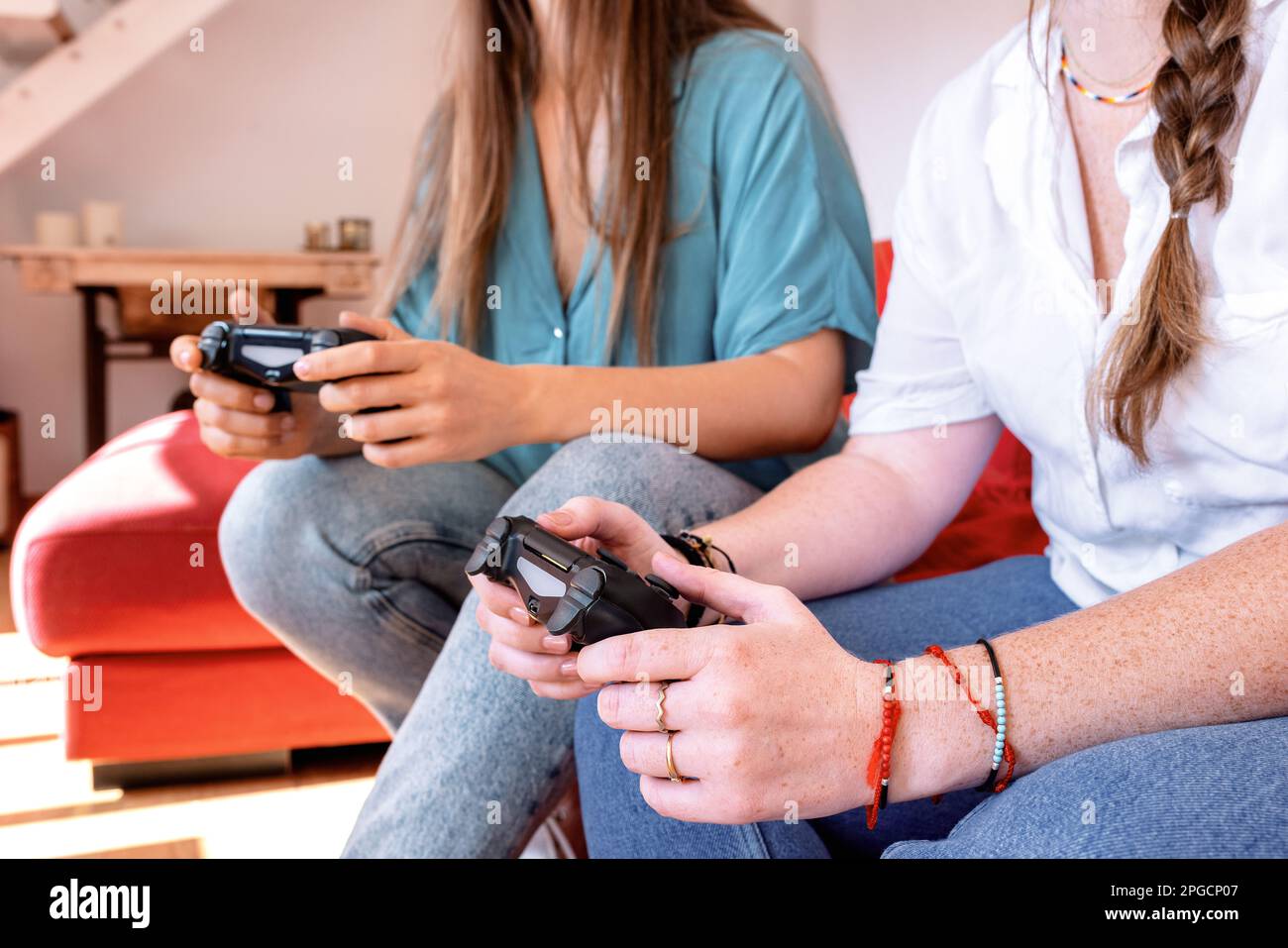 Le amiche femminili sono irriconoscibili e si rilassano su un comodo divano di colore rosso con controller per videogiochi in mano e si divertono con la dur del tempo libero Foto Stock