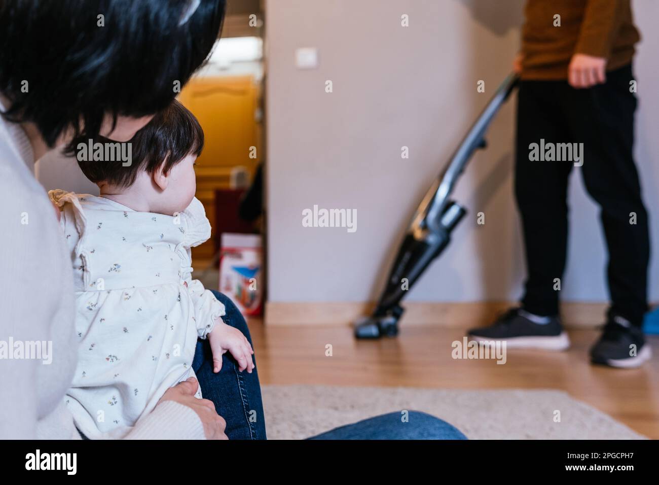 Madre irriconoscibile seduta con bambina e padre piano di pulizia con aspirapolvere in background a casa Foto Stock