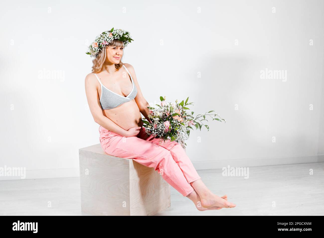 Donna incinta in corona con bouquet di fiori. Ragazza in top grigio, pantaloni rosa. Mani sul ventre nudo. Photoshoot gravidanza felice, maternità, preparazione Foto Stock
